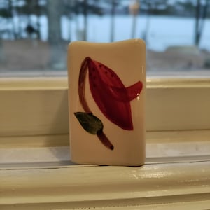 May include: A white ceramic tile with a hand-painted red flower with green leaves. The flower is stylized and has a rounded shape.