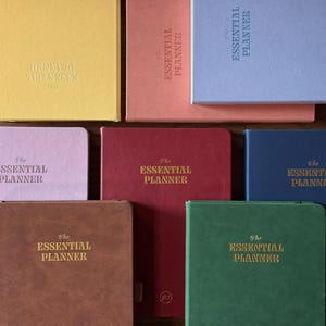 May include: A collection of eight rectangular planners in various colors including yellow, peach, light blue, light pink, dark red, dark blue, brown, and green. Each planner features the text The Essential Planner embossed on the front cover.