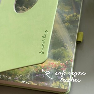 May include: A green notebook with the words "forest log" in cursive. The notebook has a cutout on the cover and is made of soft vegan leather. The inside cover features a painting of a forest scene with flowers.