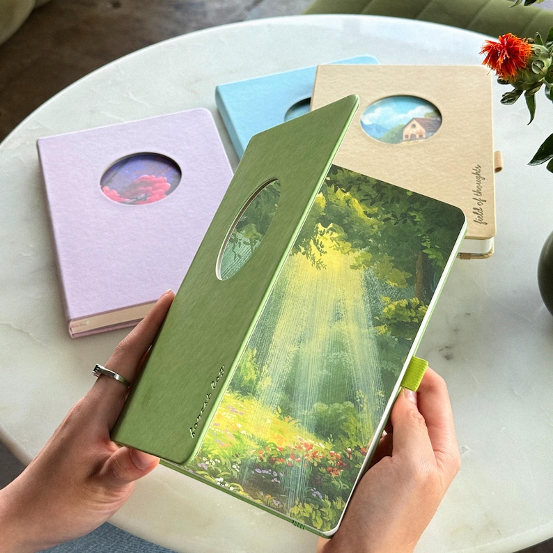 May include: A collection of hardcover journals in pastel colors, each with a circular cutout revealing a landscape image. The open green journal displays a forest scene with sunlight streaming through the trees. The text "forever calm" is visible on the cover.