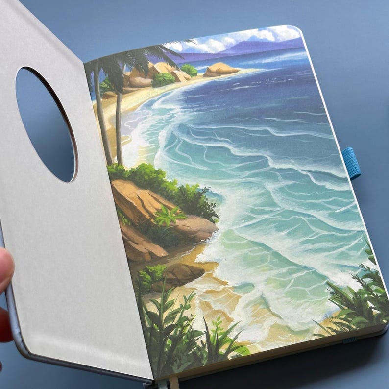 May include: Open notebook with a beach scene illustration. The artwork depicts a sandy shore, turquoise waves, palm trees, and a blue sky. The notebook has a light gray cover with a circular cutout and a blue ribbon bookmark.