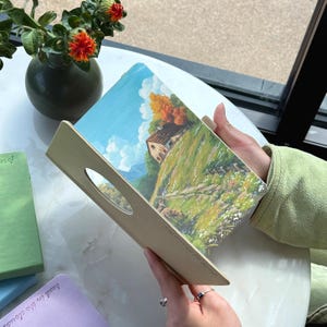 May include: A landscape painting of a house on a hill with a blue sky and clouds, displayed on a wooden tray with a handle. The tray is held by a person. Other notebooks are visible in the background.