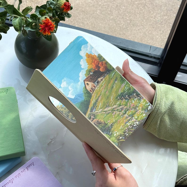 May include: A landscape painting of a house on a hill with a blue sky and clouds, displayed on a wooden tray with a handle. The tray is held by a person. Other notebooks are visible in the background.