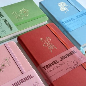 May include: A collection of floral series travel journals in various colors: green, blue, red, and pink. Each journal features a gold foil floral design and an elastic band closure. The journals are A5 size, measuring 108 x 195 mm.