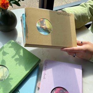 May include: A collection of notebooks in various colors, including tan, green, and purple. Each notebook features a circular cutout revealing a watercolor image. The tan notebook is held up, displaying the text "fields of thoughts."