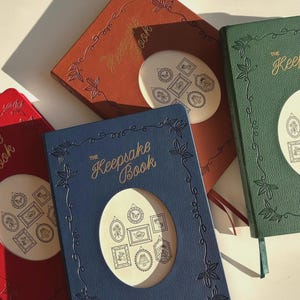 May include: Three different colored journals with the text "The Keepsake Book" on the front. The journals are blue, red, and green. Each journal has a decorative frame around the title and a cut-out window with a design of small frames inside.