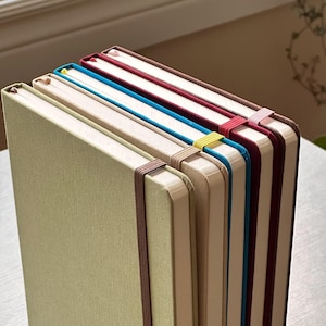 May include: A stack of four hardcover notebooks with fabric covers in sage green, beige, bright blue, and deep burgundy. Each notebook features a matching elastic closure band and a thin ribbon bookmark, arranged vertically in a neat, upright row.