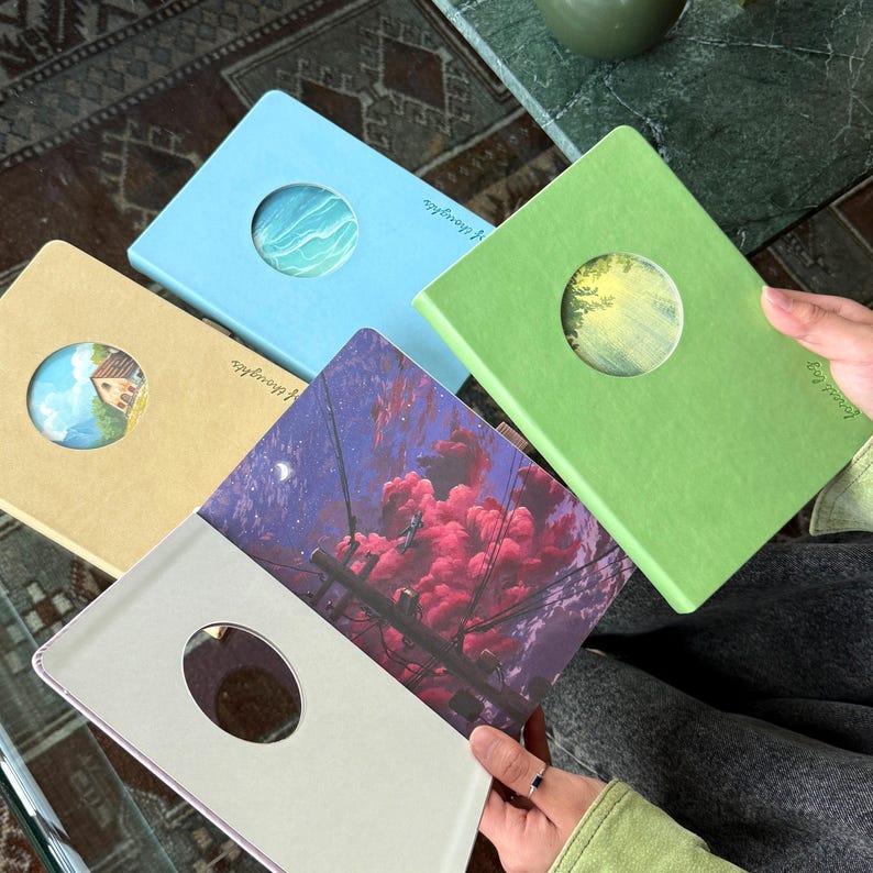 May include: A collection of notebooks in various colors, including light blue, green, and tan. Each notebook features a circular cutout revealing a unique illustration. The open notebook displays a vibrant image of a night sky with pink clouds.