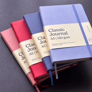 May include: Four A5 journals with textured covers in light purple, dark blue, red, and pink. Each journal features a cream-colored paper band printed with "Classic Journal A5 | 150 gsm", a matching elastic closure strap, and a ribbon bookmark.