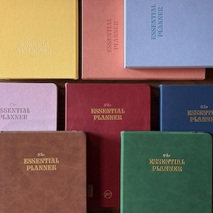 May include: A collection of eight rectangular planners in various colors including yellow, peach, light blue, light pink, dark red, dark blue, brown, and green. Each planner features the text The Essential Planner embossed on the front cover.