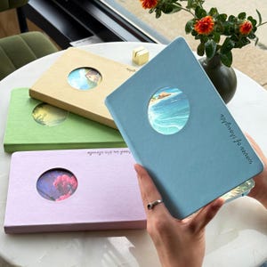 May include: Four journals in pastel shades: light blue, green, beige, and pink. Each journal features a circular aperture showcasing a watercolour image. The light blue journal is held open, revealing a beach scene. Text reads "waves of thoughts".