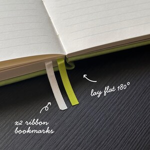 May include: Open notebook with lined pages and two ribbon bookmarks. One bookmark is white, the other is lime green. The notebook is open to a 180-degree angle, as indicated by the text in the image.