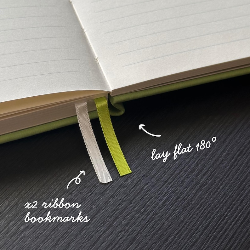 May include: Open notebook with lined pages and two ribbon bookmarks. One bookmark is white, the other is lime green. The notebook is open to a 180-degree angle, as indicated by the text in the image.