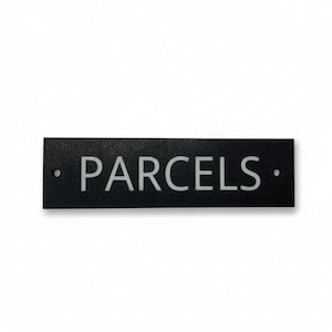 May include: A rectangular black sign with the word PARCELS printed in white capital letters. The sign features a textured surface and includes two small circular mounting holes, one on the far left and one on the far right side of the plate.
