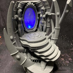 May include: A grey, futuristic sculpture with a central oval portal emitting blue light. Two humanoid figures flank the portal. The sculpture has a tiered base and ornate, curved supports, creating a sci-fi or fantasy aesthetic.