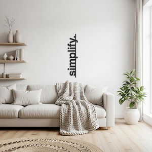 Puede incluir: Un salón moderno con un sofá blanco, cojines y una gruesa manta de punto. Una decoración de pared muestra la palabra "simplify". Una planta en maceta y una alfombra redonda completan el diseño minimalista.