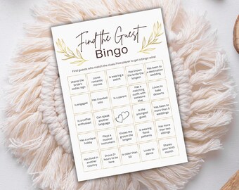 以下が含まれることがあります： 「Find The Guest Bingo」と金色の文字が書かれた白いビンゴゲームカード。カードには、「花嫁の星座を共有する」など、ゲストが一致させるための手がかりのグリッドがあります。クリーム色のマクラメコースターの上にあります。