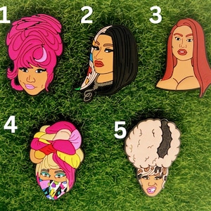 May include: Five colorful enamel pins featuring different women with unique hairstyles. The pins are on a green background.