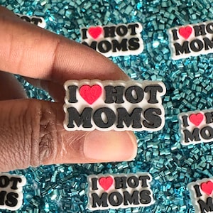 May include: A close-up of a white, red, and black charm with the text "I love hot moms" and a red heart. The charm is surrounded by turquoise glitter.