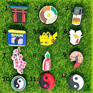May include: Twelve colourful charms with various designs, including a Japanese gate, a bowl of rice, a can of Spam, a Pokemon character, a cherry blossom branch, a bottle of hot sauce, a shrimp, and a yin yang symbol.