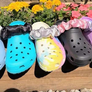 May include: Five pairs of colorful clogs with matching scrunchies. The clogs are in light blue, turquoise, tie-dye, black, and lavender. Each clog has a scrunchie in a coordinating color. The clogs are displayed on a wooden surface.