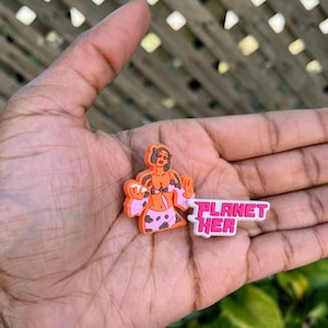 May include: An orange and pink shoe charm featuring a cartoon figure and the text "PLANET HER" in pink and white. The charm has a pink and white spotted pattern on the figure's clothing.