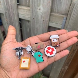 May include: A set of seven colorful nurse-themed shoe charms. The charms include a stethoscope, a bandage, a pill bottle, a medical symbol, a red cross, a clipboard with a red cross, and a green scrub top with a red heart.