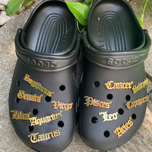 May include: Black Crocs with gold zodiac charms attached to the shoe. The charms include the signs: Sagittarius, Gemini, Virgo, Libra, Aquarius, Taurus, Cancer, Scorpio, Capricorn, Pisces, Leo, and Aries.