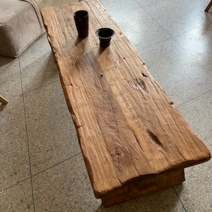 May include: A rustic, rectangular wooden coffee table with a natural, unfinished look. The table has a rough-hewn surface and a warm, brown color. Two small, dark-colored decorative cups sit on the table. The table is approximately 60 inches long.