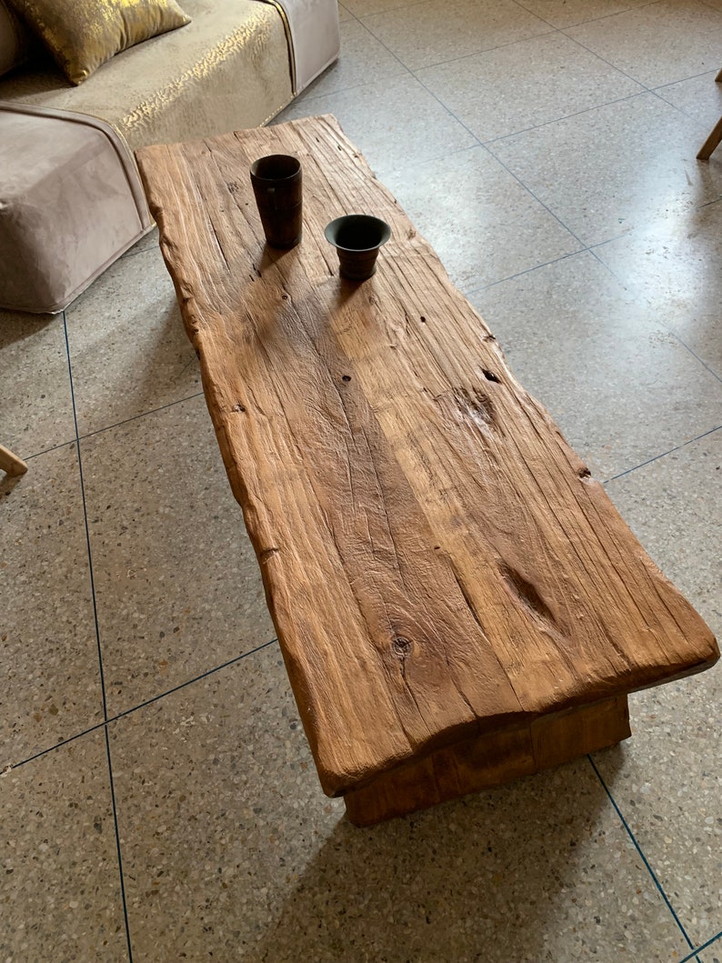 May include: A rustic, rectangular wooden coffee table with a natural, unfinished look. The table has a rough-hewn surface and a warm, brown color. Two small, dark-colored decorative cups sit on the table. The table is approximately 60 inches long.