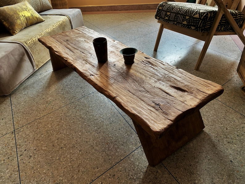 May include: A rustic, rectangular coffee table made of weathered wood. The table has a natural edge and sits on two angled supports. Two dark, cylindrical containers are on the table's surface. The table is in a living room setting.