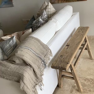 May include: A rustic, weathered wooden bench with a rectangular seat and angled legs stands beside a white sofa. A beige throw blanket with dark blue stripes and fringe rests on the sofa. Decorative pillows in various patterns add texture and color.