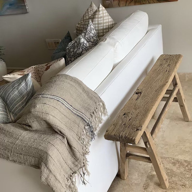 May include: A rustic, weathered wooden bench with a rectangular seat and angled legs stands beside a white sofa. A beige throw blanket with dark blue stripes and fringe rests on the sofa. Decorative pillows in various patterns add texture and color.