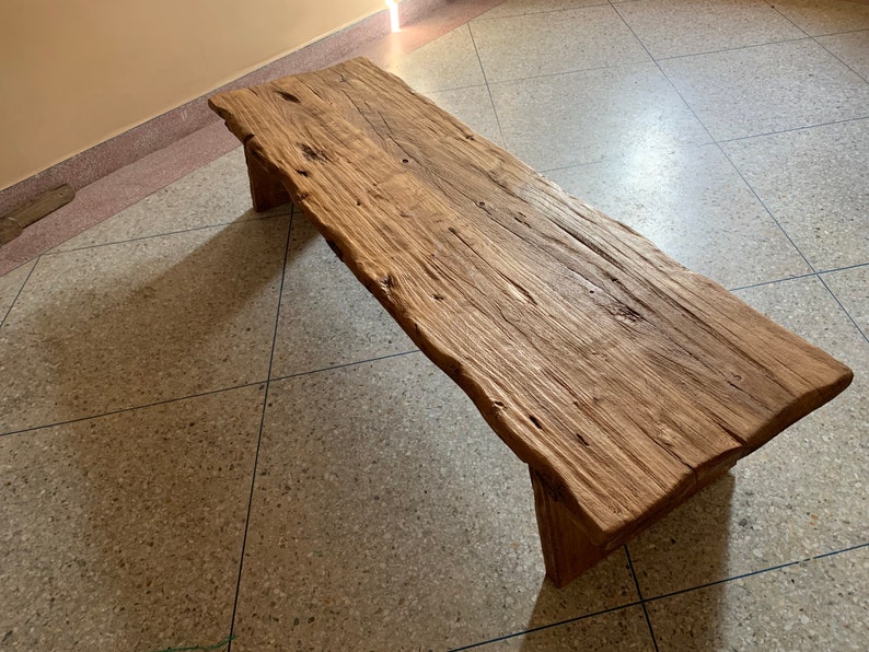 May include: A rustic wooden bench with a natural, unfinished look. The bench is made from a single, thick slab of wood, supported by two sturdy legs. The wood has a warm, brown tone with visible grain and knots. The bench is approximately 60 inches long.