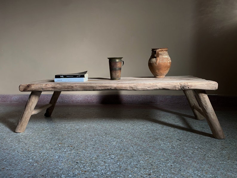 K&ouml;nnte beinhalten: Ein rustikaler, niedriger Holztisch mit angewinkelten Beinen. Auf dem Tisch befinden sich ein Stapel B&uuml;cher, ein Messingbecher und eine Terrakotta-Vase. Die nat&uuml;rliche Holzmaserung des Tisches und die gealterte Keramik deuten auf eine Vintage- oder antike &Auml;sthetik hin. Der Tisch ist etwa 150 cm lang.