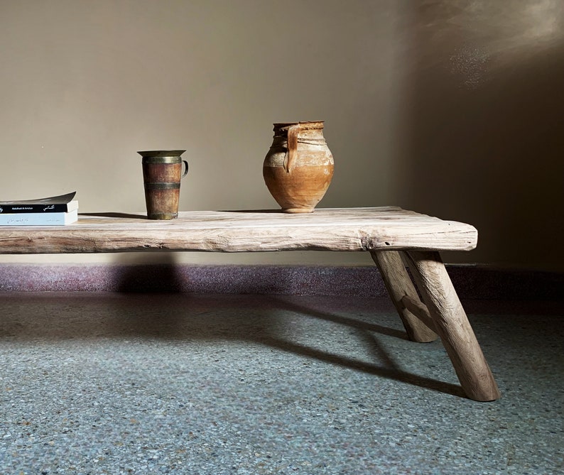 K&ouml;nnte beinhalten: Ein rustikaler Holztisch mit nat&uuml;rlicher, unbehandelter Optik. Er hat ein verwittertes Aussehen und stabile, angewinkelte Beine. Auf dem Tisch befinden sich ein Vintage-Messingbecher, ein Terrakotta-Krug und ein Stapel B&uuml;cher. Die Szene wird von weichem, nat&uuml;rlichem Licht durchflutet.