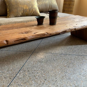 May include: A rustic, low-profile wooden coffee table with a natural edge. The table is made of light brown wood and has a simple, geometric base. Two small, dark brown containers sit on the table's surface.
