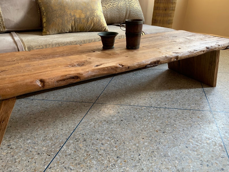 May include: A rustic, low-profile wooden coffee table with a natural edge. The table is made of light brown wood and has a simple, geometric base. Two small, dark brown containers sit on the table's surface.