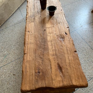 May include: A rectangular, rustic coffee table made of light brown wood. The table has a natural edge and visible wood grain. Two dark brown, cylindrical containers sit on the table's surface. The table is approximately 60 inches long.