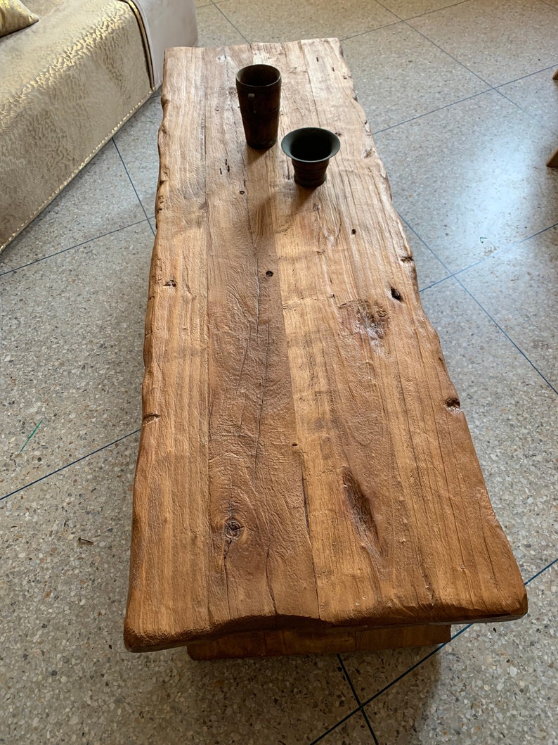 May include: A rectangular, rustic coffee table made of light brown wood. The table has a natural edge and visible wood grain. Two dark brown, cylindrical containers sit on the table's surface. The table is approximately 60 inches long.