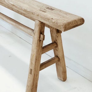 May include: A light-colored wooden bench with a rectangular seat and angled legs. The bench is made of weathered wood, showcasing natural grain and knots. The legs are connected by horizontal supports, creating a sturdy structure.