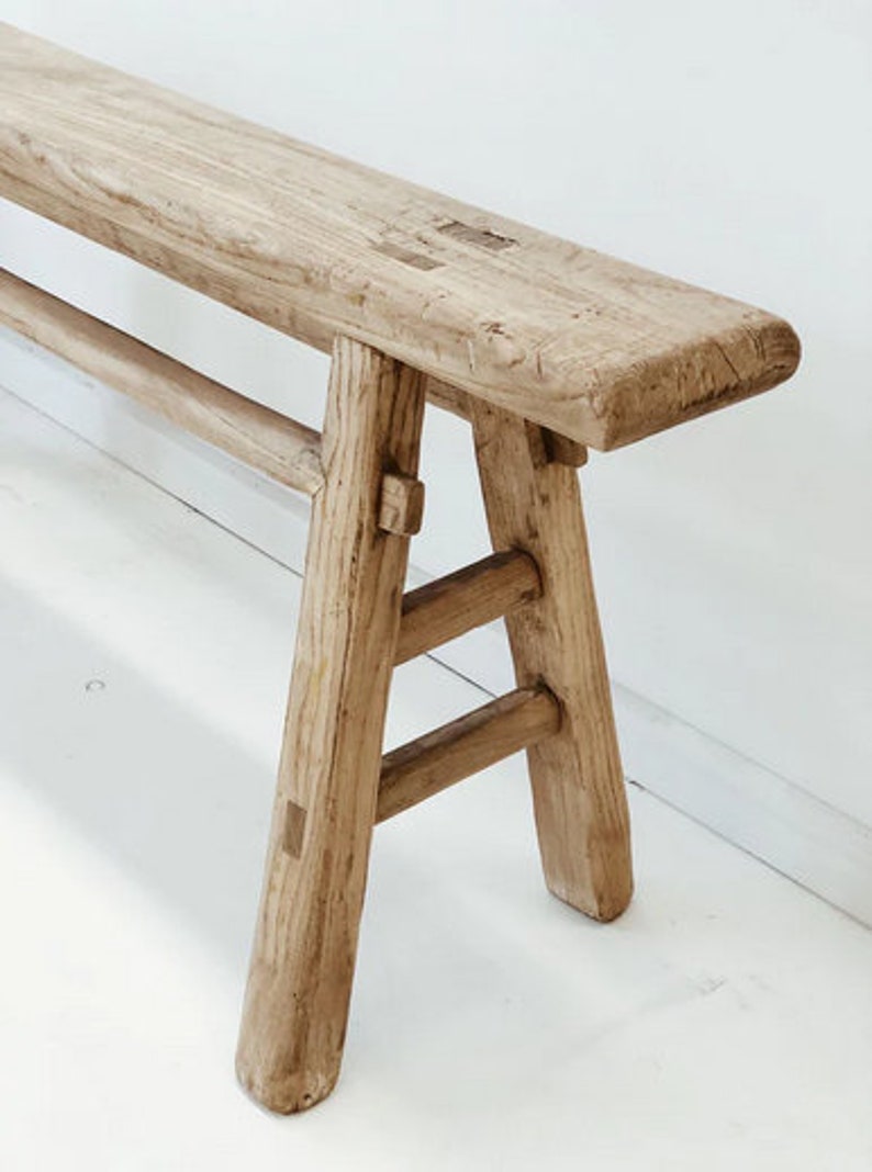 May include: A light-colored wooden bench with a rectangular seat and angled legs. The bench is made of weathered wood, showcasing natural grain and knots. The legs are connected by horizontal supports, creating a sturdy structure.