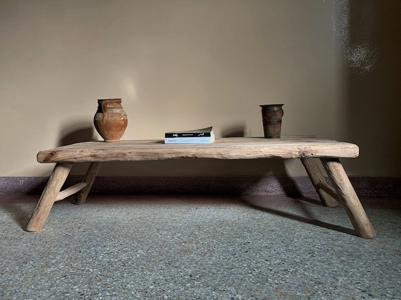 K&ouml;nnte beinhalten: Ein rustikaler, niedriger Holztisch mit nat&uuml;rlicher Oberfl&auml;che, getragen von angewinkelten Beinen. Zwei antike Gef&auml;&szlig;e und ein Buch liegen auf der Tischoberfl&auml;che. Der Tisch ist etwa 152 cm lang.