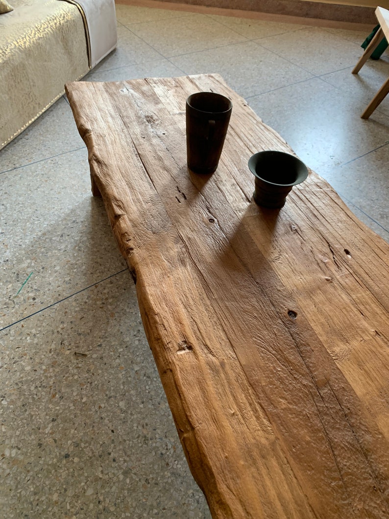 May include: A rustic, rectangular wooden coffee table with a natural edge. Two dark brown containers sit on the table. The table's surface shows the natural grain and knots of the wood. The table is in a room with a tiled floor.