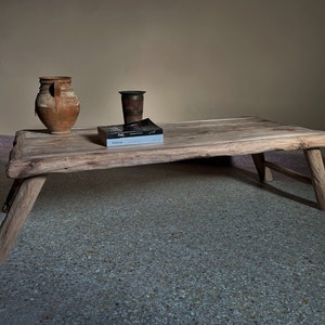 K&ouml;nnte beinhalten: Ein rustikaler, niedriger Couchtisch aus Holz mit einem nat&uuml;rlichen, unbehandelten Aussehen. Der Tisch hat drei abgewinkelte Beine und eine rechteckige Platte. Ein verwitterter Tonkrug, eine kleine Metallvase und ein Buch liegen auf dem Tisch. Der Hintergrund ist neutral.
