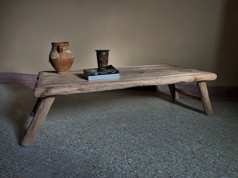 K&ouml;nnte beinhalten: Ein rustikaler, niedriger Couchtisch aus Holz mit einem nat&uuml;rlichen, unbehandelten Aussehen. Der Tisch hat drei abgewinkelte Beine und eine rechteckige Platte. Ein verwitterter Tonkrug, eine kleine Metallvase und ein Buch liegen auf dem Tisch. Der Hintergrund ist neutral.