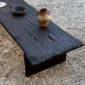 Peut inclure: Une table en bois teinté foncé avec un bord naturel. Un petit bol foncé, un petit livre et un vase beige à deux anses sont posés sur la table. La table est faite d'une seule pièce de bois épaisse.