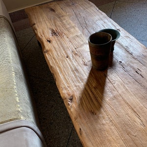 May include: A rustic, light brown wooden coffee table with a natural edge. Two small, dark brown wooden cups sit on the table. The table has visible wood grain and knots, and is next to a light-colored sofa.