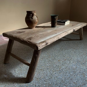 K&ouml;nnte beinhalten: Ein rustikaler, niedriger Couchtisch aus Holz mit nat&uuml;rlicher, unbehandelter Optik. Der Tisch hat vier angewinkelte Beine und eine Baumkante. Eine braune Keramikvase, eine dunkle Tasse und ein Buch liegen auf der Tischoberfl&auml;che. Der Tisch ist ca. 122 cm lang.