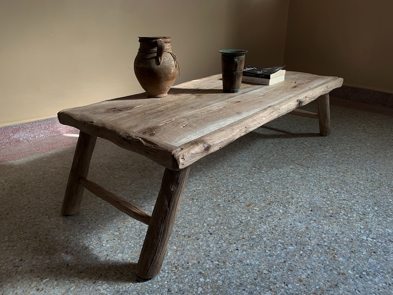 K&ouml;nnte beinhalten: Ein rustikaler, niedriger Couchtisch aus Holz mit nat&uuml;rlicher, unbehandelter Optik. Der Tisch hat vier angewinkelte Beine und eine Baumkante. Eine braune Keramikvase, eine dunkle Tasse und ein Buch liegen auf der Tischoberfl&auml;che. Der Tisch ist ca. 122 cm lang.
